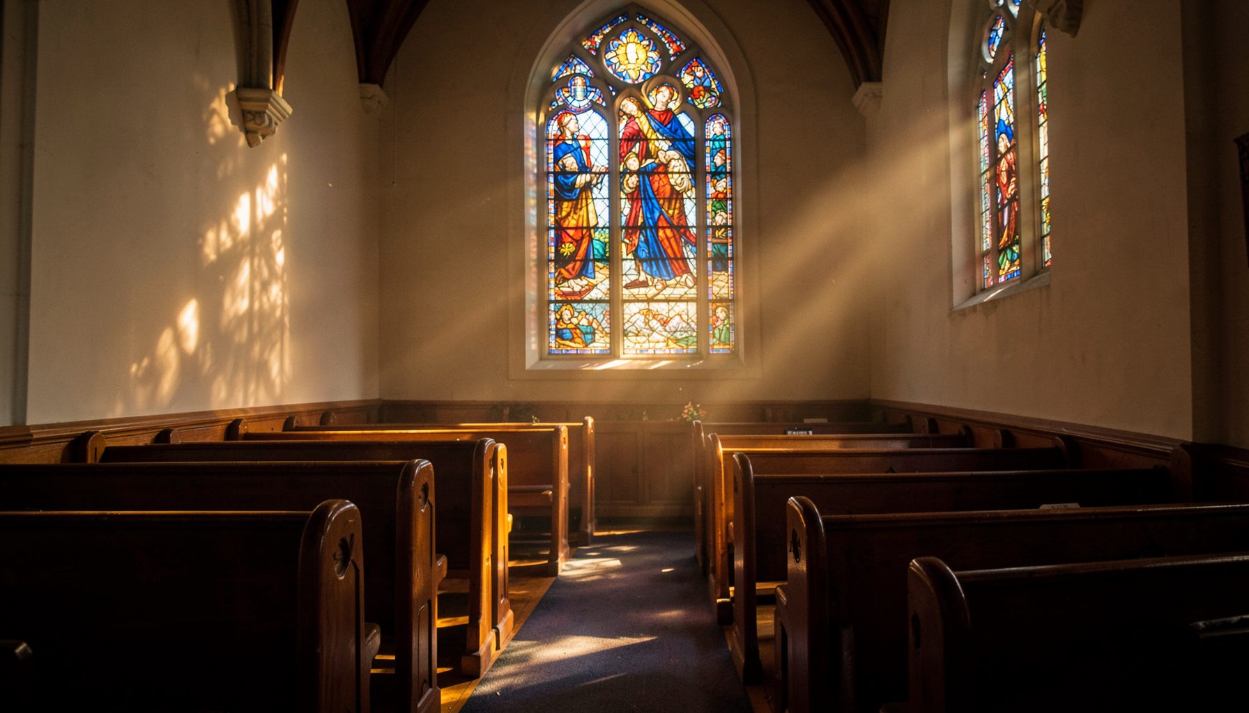 Sunlight streaming through a stained-glass window in a quiet church sanctuary, casting warm amber and deep blue tones across wooden pews.