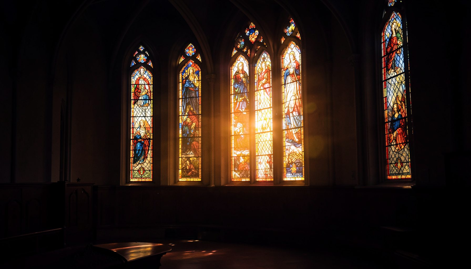 Warm golden light filtering through a row of tall stained-glass windows in a quiet sanctuary, with deep navy and amber tones